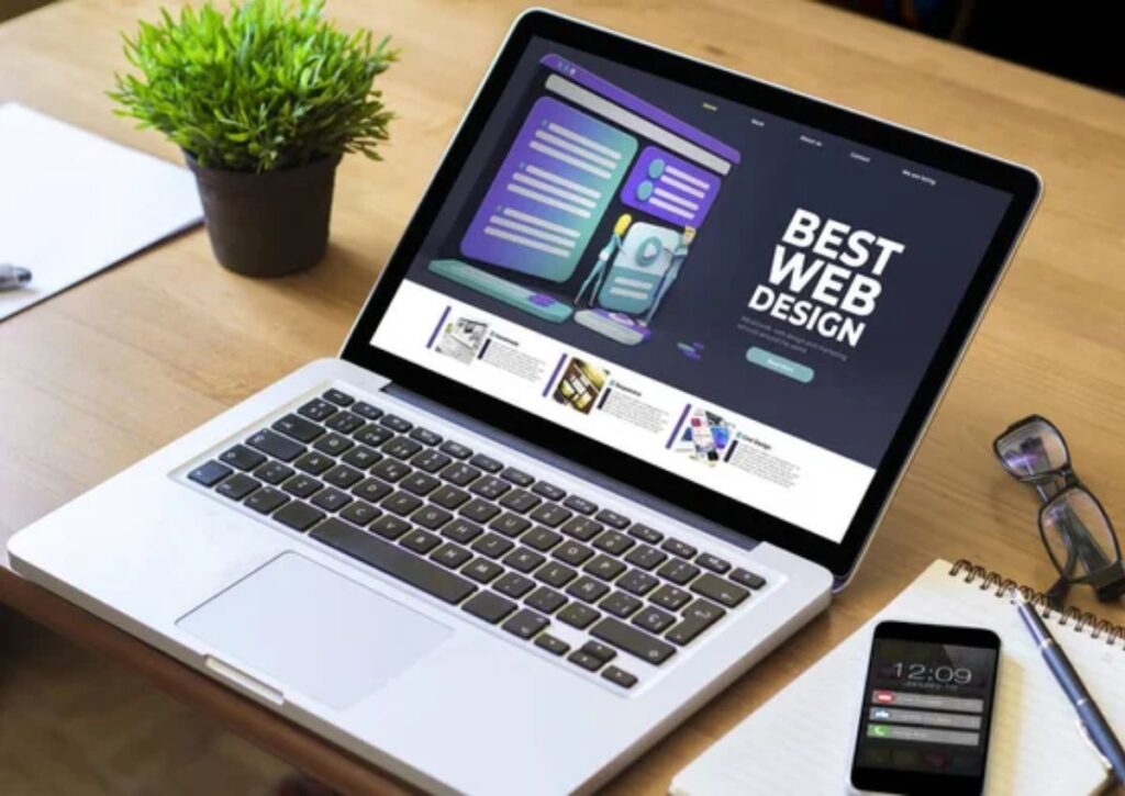Simply The Best Digital Marketing photo of laptop displaying "Best Web Design" title on a responsive web design page on a wooden table with a green plant, a mobile phone, glasses, and a notepad with a pen next to it