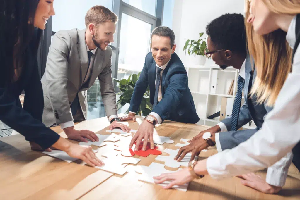 simply the best digital marketing team photo of people standing around at light brown desk piecing a puzzle together.