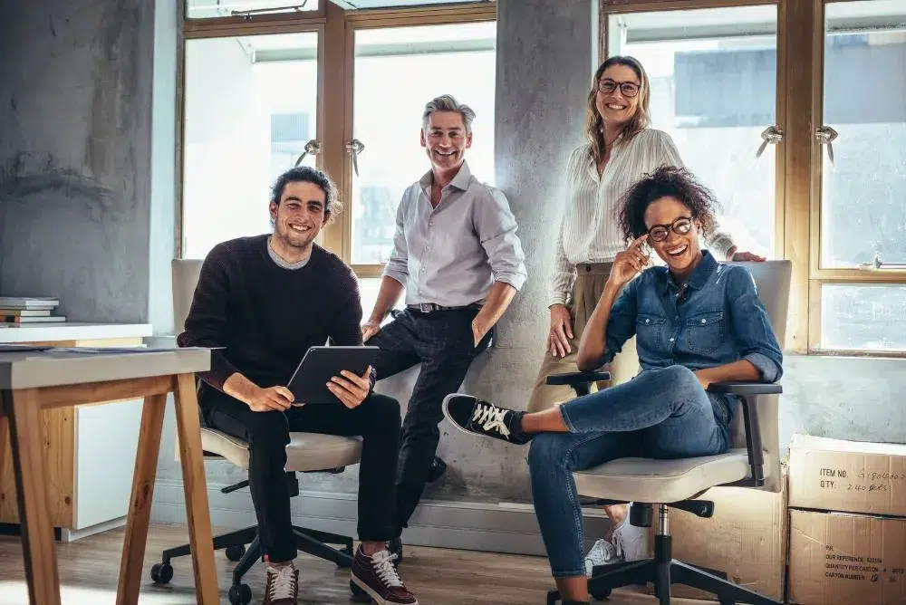 Four cheerful professionals in a modern office setting, with one man holding a tablet and the other three individuals casually posing around a desk.